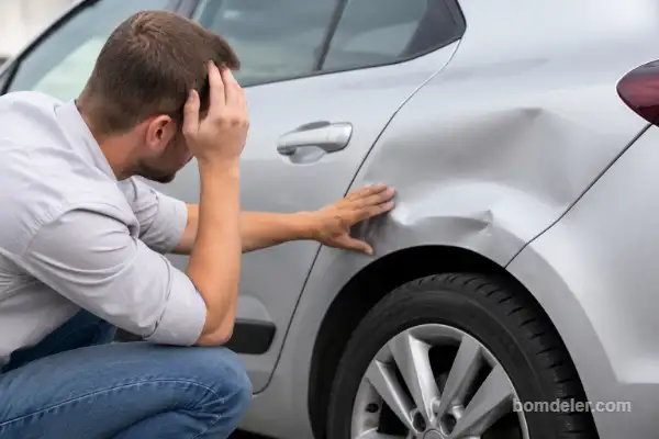 Motorista preocupado analisando amassado na lateral de um carro prata após colisão